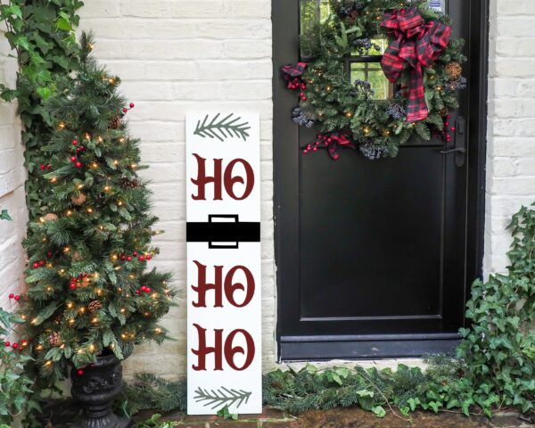 A black door features a 11x48 "Ho, Ho, Ho" white sign with a belt design, a small lit Christmas tree, a festive wreath, and surrounding greenery. - Spruced Studio Upscale Workshop