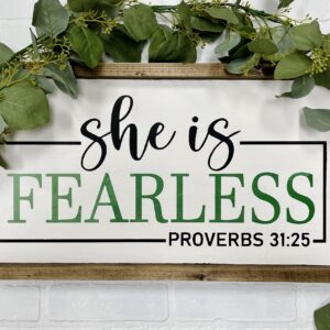 The 13x24 She is Fearless sign features green leaves and the text "She is fearless. Proverbs 31:25" in black and green on a white background. - Spruced Studio Upscale Workshop