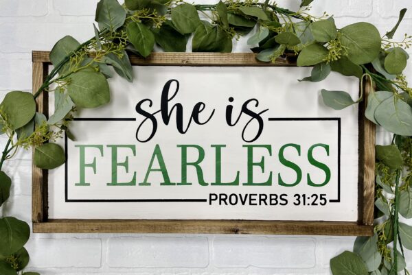 The 13x24 She is Fearless sign features green leaves and the text "She is fearless. Proverbs 31:25" in black and green on a white background. - Spruced Studio Upscale Workshop