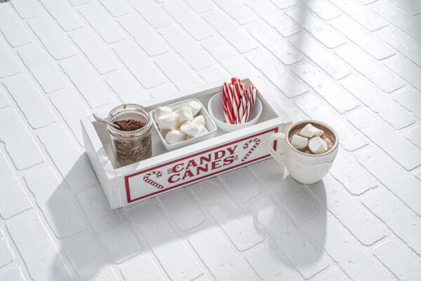 A Sm Box Candy Canes tray holds a jar of cocoa mix, marshmallows, and candy canes. A mug of hot chocolate with marshmallows sits beside the tray on a white brick surface. - Spruced Studio Upscale Workshop