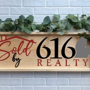 A 12x32 "Sold By 616" wooden sign with greenery features a house roof graphic and Michigan state outline, displayed against a white brick wall. - Spruced Studio Upscale Workshop