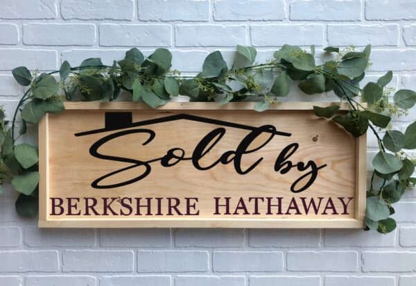 A 12x32 "Sold By BHHS" wooden sign with greenery, featuring black and maroon lettering, is mounted on a white brick wall. - Spruced Studio Upscale Workshop