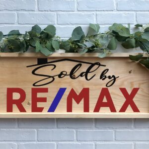 A 12x32 "Sold By REMAX" wooden sign with green leaves is displayed against a white brick wall. - Spruced Studio Upscale Workshop