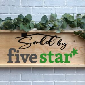 A 12x32 Sold By FiveStar wooden sign, featuring green foliage and "Sold by five star*" in black and green lettering, is displayed against a white brick wall. - Spruced Studio Upscale Workshop