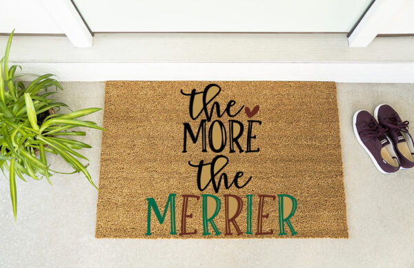 The More The Merrier doormat features black and green lettering, displayed at a doorway with a green plant and maroon shoes nearby. - Spruced Studio Upscale Workshop
