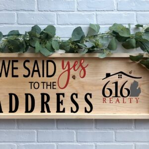 The 12x32 Said Yes to the Address - 616 features a wooden sign with greenery, displaying "We said yes to the address 616 Realty" in black and red text against a white brick wall. - Spruced Studio Upscale Workshop