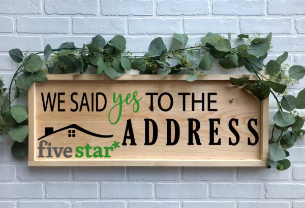 The 12x32 Said Yes to the Address - FiveStar wooden sign with greenery reads "We said yes to the address five star*" and is displayed against a white brick wall background. - Spruced Studio Upscale Workshop