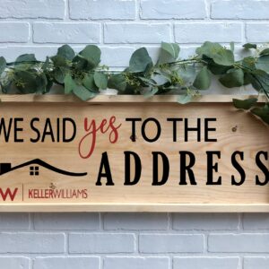 The 12x32 "Said Yes to the Address - Keller Williams" wooden sign, featuring green leaves and the Keller Williams logo, is displayed on a white brick wall. - Spruced Studio Upscale Workshop