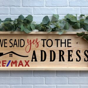 The 12x32 Said Yes to the Address - REMAX is a wooden sign with greenery, featuring "We said yes to the address," the RE/MAX logo, and a house graphic, displayed on a white brick wall. - Spruced Studio Upscale Workshop