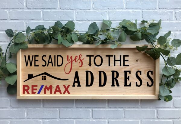 The 12x32 Said Yes to the Address - REMAX is a wooden sign with greenery, featuring "We said yes to the address," the RE/MAX logo, and a house graphic, displayed on a white brick wall. - Spruced Studio Upscale Workshop