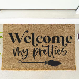 The "Welcome My Pretties" doormat, featuring a broom illustration, is placed at the door alongside a potted plant and maroon shoes. - Spruced Studio Upscale Workshop