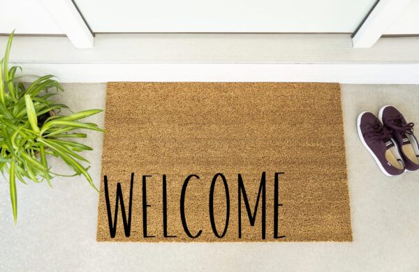 The Welcome 1 doormat, featuring "WELCOME" in black letters, is placed by a white door with a green potted plant and maroon shoes nearby. - Spruced Studio Upscale Workshop