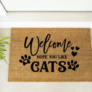 The Welcome Hope You Like Cats doormat features black paw prints and is shown in front of a door beside a plant and maroon shoes. - Spruced Studio Upscale Workshop