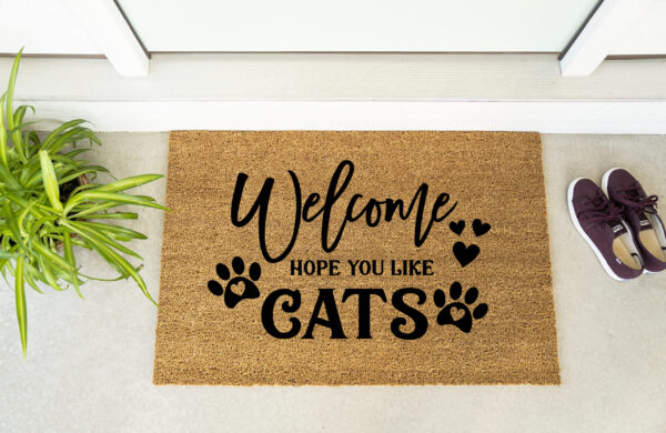 The Welcome Hope You Like Cats doormat features black paw prints and is shown in front of a door beside a plant and maroon shoes. - Spruced Studio Upscale Workshop