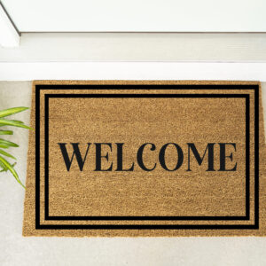 The Welcome w/ Frame doormat sits in front of a white door, accompanied by a green potted plant and maroon shoes. - Spruced Studio Upscale Workshop