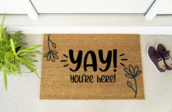 The "Yay You're Here" doormat features black lettering and looks welcoming by the door, with a plant on the left and maroon shoes on the right. - Spruced Studio Upscale Workshop