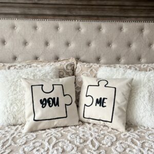 Two "You + Me" decorative pillows with puzzle piece designs and the words "YOU" and "ME" sit on a bed with a beige tufted headboard and textured bedding. - Spruced Studio Upscale Workshop