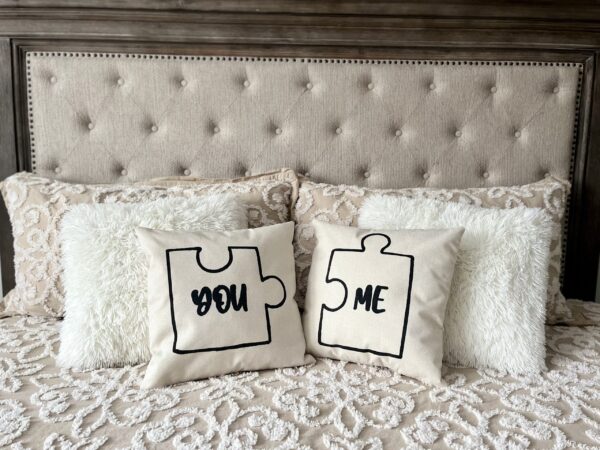 Two "You + Me" decorative pillows with puzzle piece designs and the words "YOU" and "ME" sit on a bed with a beige tufted headboard and textured bedding. - Spruced Studio Upscale Workshop