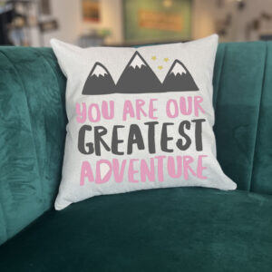 The "You are Our Greatest Adventure" pillow features a mountain graphic and pink and gray text, displayed on a green velvet couch. - Spruced Studio Upscale Workshop