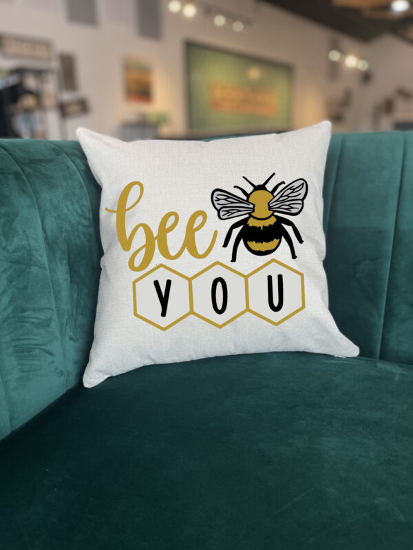 The Bee You throw pillow is white with "bee you" text, a bee illustration, and honeycomb shapes, displayed on a green couch. - Spruced Studio Upscale Workshop