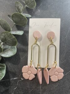 A pair of pink and gold dangle earrings with floral and leaf designs on a card labeled "One Twelve," next to green leaves on a dark surface. - Spruced Studio Upscale Workshop