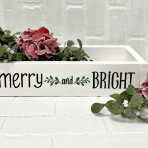 The Small Box - Merry & Bright is a white wooden box featuring "merry and BRIGHT" on the side, filled with artificial red flowers and green leaves, placed on a white brick surface. - Spruced Studio Upscale Workshop