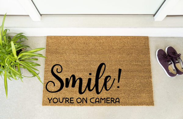 The Smile! You're on Camera doormat is placed at a doorway beside a plant and maroon shoes. - Spruced Studio Upscale Workshop