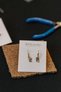 A pair of gold earrings displayed on a white card, positioned on a cork mat, with blue-handled pliers in the background. - Spruced Studio Upscale Workshop