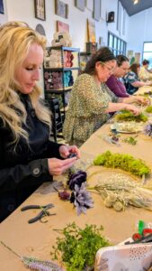 Several women sit at a long table in a craft studio, making floral arrangements with various dried plants and flowers. Craft tools and supplies are spread out on the table. - Spruced Studio Upscale Workshop