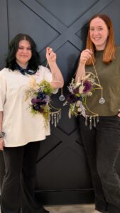 Two women stand side by side, each holding a decorative hanging ornament with flowers, greenery, crystals, and beads, against a dark paneled wall. - Spruced Studio Upscale Workshop