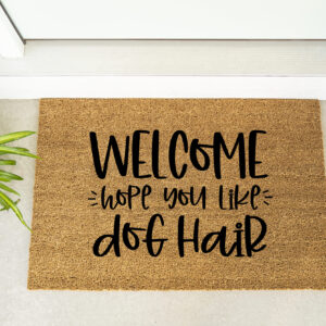 The Welcome Hope You Like Dog Hair doormat is placed in front of a door, with a plant and maroon shoes nearby. - Spruced Studio Upscale Workshop