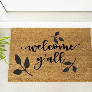The "Welcome Y'all" doormat featuring leaf designs is placed by a white door, accompanied by a green potted plant and maroon shoes. - Spruced Studio Upscale Workshop