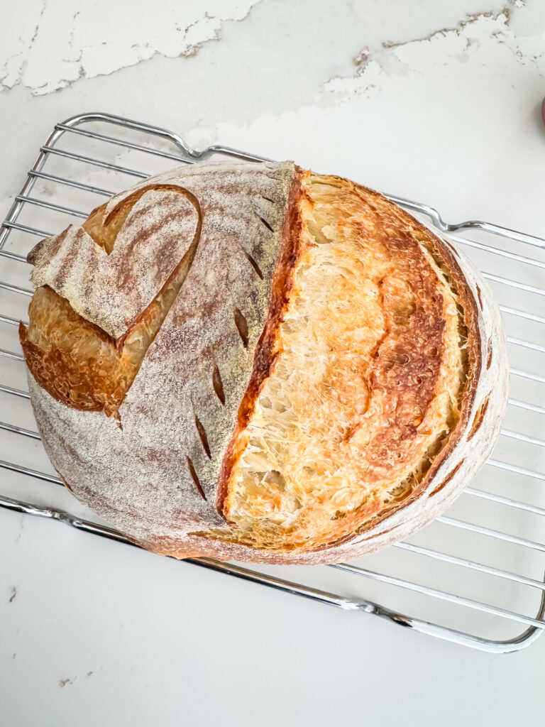 A round loaf of sourdough bread with a golden crust rests on a metal cooling rack atop a white countertop. - Spruced Studio Upscale Workshop