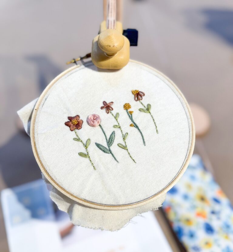 An embroidery hoop holds fabric with hand-stitched flowers, surrounded by blurred objects including a floral pouch and a booklet. - Spruced Studio Upscale Workshop