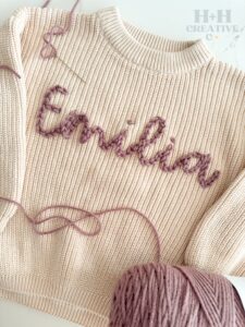 A cream-colored knit sweater with the name "Emilia" embroidered in pink yarn, shown with a ball of matching yarn and loose threads. - Spruced Studio Upscale Workshop