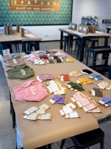 Table with embroidery threads organized by color, embroidery hoops, and fabric pieces labeled “Aloha” and “Ohana” in a craft workshop setting. - Spruced Studio Upscale Workshop