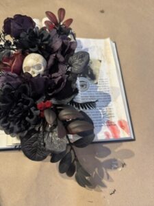 Open book with dark artificial flowers, leaves, and a small skull placed on top, alongside red stains resembling blood on the book’s pages. - Spruced Studio Upscale Workshop