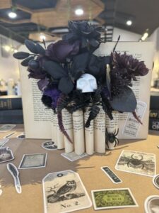 A bouquet of dark flowers with a skull ornament is displayed in an open book, surrounded by vintage-themed stickers and Halloween decorations on a brown table. - Spruced Studio Upscale Workshop