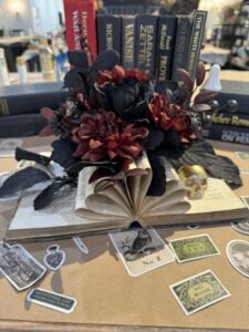 An open book with artificial red and black flowers on it is displayed on a table, surrounded by stickers and books. - Spruced Studio Upscale Workshop