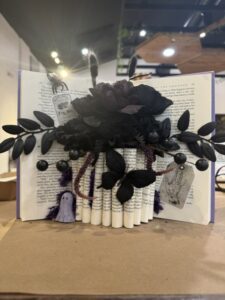 An open book stands upright, decorated with black artificial flowers, leaves, fake berries, tarot cards, and a small purple ghost figurine on a brown surface. - Spruced Studio Upscale Workshop