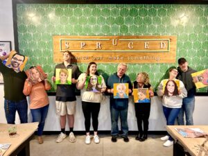 Seven people stand in a row holding painted portraits of themselves in front of a green patterned wall and a sign that reads “Spruced. Branch out, make something.”. - Spruced Studio Upscale Workshop