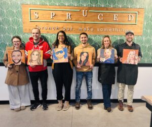 Six people stand in a row holding painted portraits in front of a "Spruced" sign with the tagline "branch out, make something. - Spruced Studio Upscale Workshop