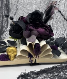 An open book with pages folded into a heart shape, topped with black and purple artificial flowers, next to a small gold skull and purple moss, all set against a black netted backdrop. - Spruced Studio Upscale Workshop