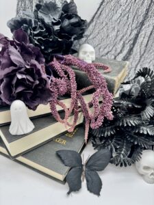 A Halloween-themed still life with black books, purple and black flowers, faux skulls, a ghost figurine, dark leaves, a purple garland, and a black butterfly against a netted backdrop. - Spruced Studio Upscale Workshop