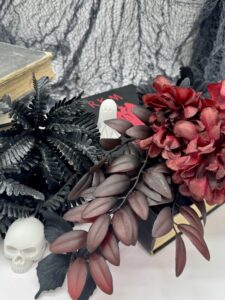 A skull, a black plant, red artificial flowers, and a small ghost figurine are arranged on and around old books against a gray cobweb backdrop. - Spruced Studio Upscale Workshop