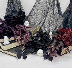 Three stacks of books decorated with artificial black and red flowers, small skull and ghost figurines, black butterflies, and gauzy black fabric in the background. - Spruced Studio Upscale Workshop