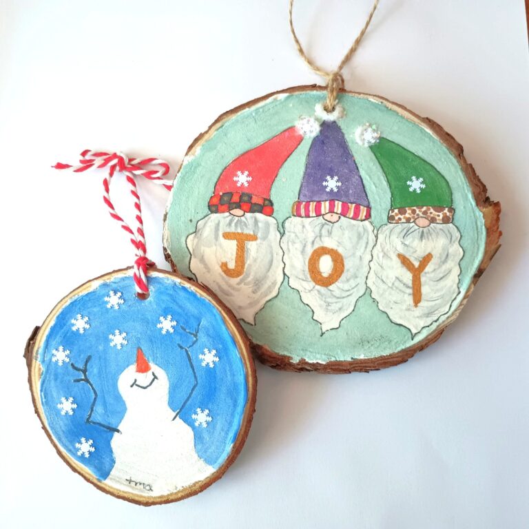 Two wooden ornaments: one with three gnomes and "JOY" painted on it, the other with a snowman and snowflakes on a blue background. - Spruced Studio Upscale Workshop