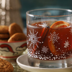 A glass of dark beverage with orange slices and a cinnamon stick, decorated with snowflake patterns, sits on a white saucer next to a bowl of cookies. - Spruced Studio Upscale Workshop
