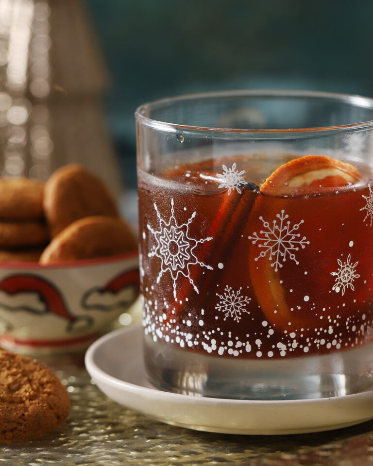 A glass of dark beverage with orange slices and a cinnamon stick, decorated with snowflake patterns, sits on a white saucer next to a bowl of cookies. - Spruced Studio Upscale Workshop
