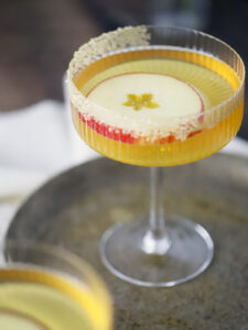 A coupe glass filled with a yellow cocktail, garnished with a thin apple slice and a crystallized sugar rim, placed on a round tray. - Spruced Studio Upscale Workshop